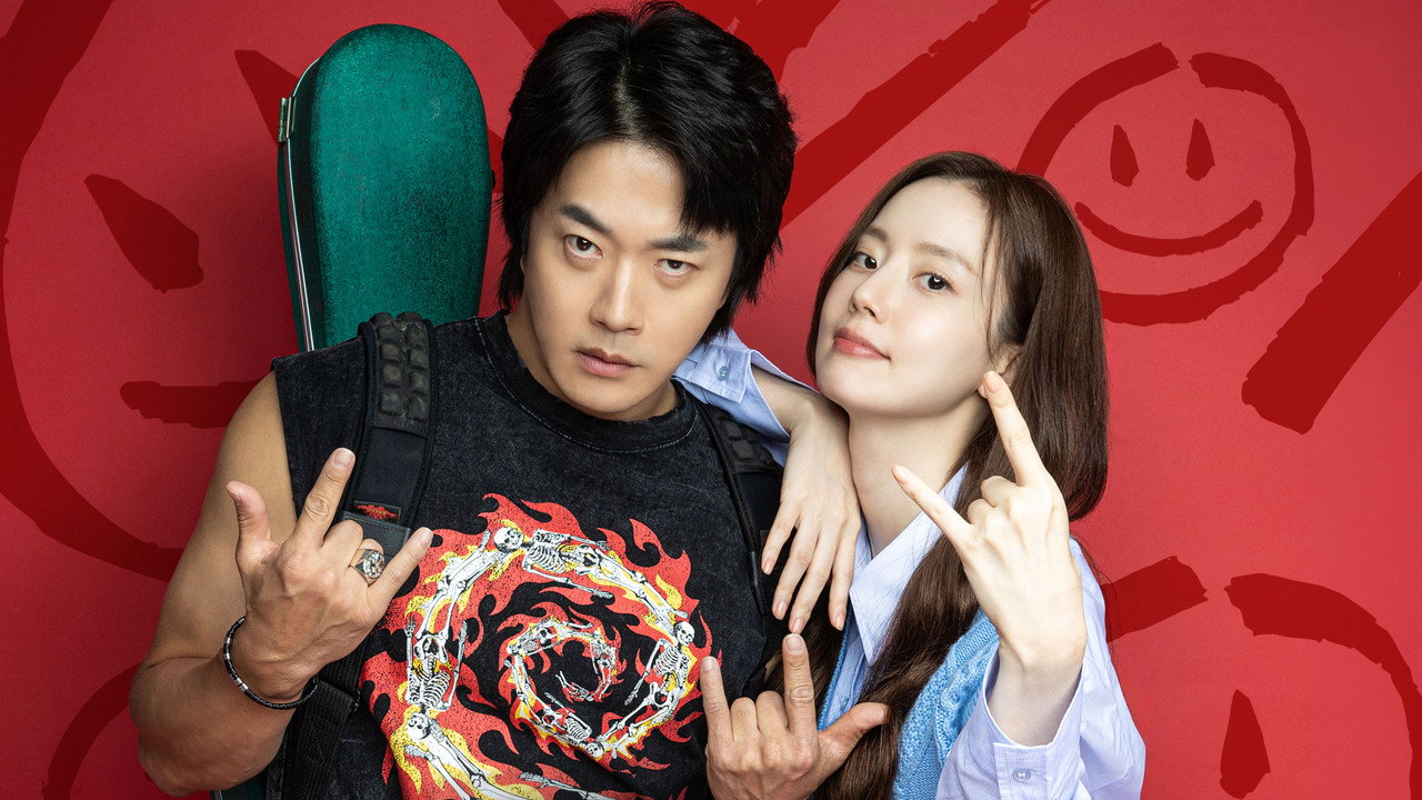 빨간 배경 앞에서 기타 케이스를 멘 남자와 손가락 하트를 든 여자가 나란히 선 하트맨 홍보 스틸
