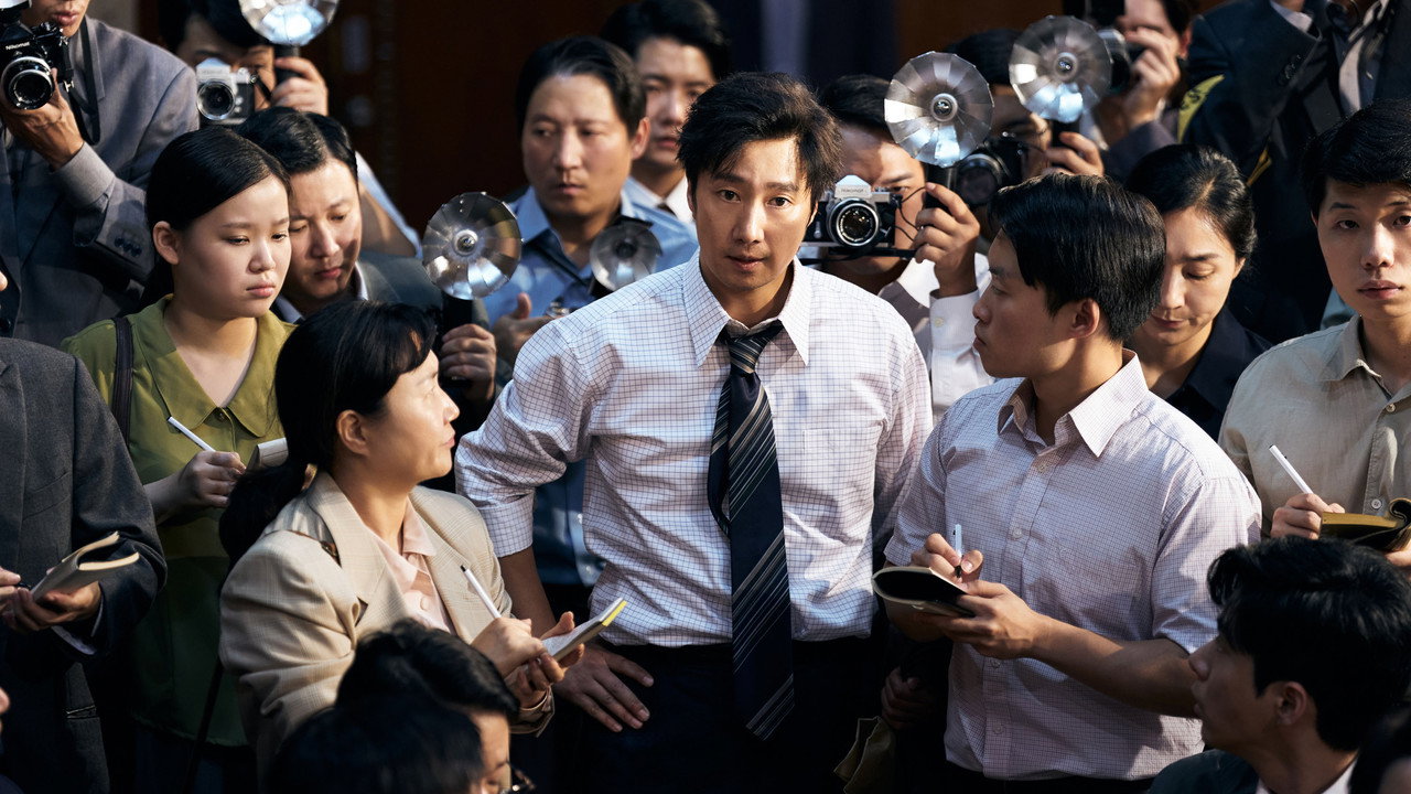 암살자(들) 스틸, 박해일이 수첩과 카메라 플래시에 둘러싸인 취재 인파 한가운데 서 있는 장면